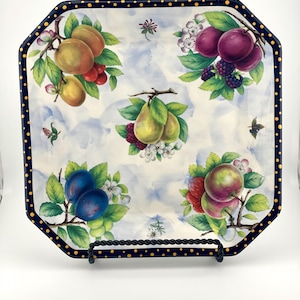 May include: A square ceramic plate with a dark blue border featuring yellow dots. The center displays a light blue watercolor-style background decorated with clusters of various fruits including pears, plums, apples, and berries with green leaves.