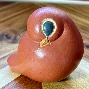May include: A smooth, terracotta-colored ceramic bird figurine with a rounded, abstract shape. It features a single dark blue eye outlined in gold, and a small, pointed gold beak. The surface has a matte finish and a simple, minimalist form.
