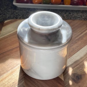 May include: A cylindrical white marble butter keeper with a smooth, polished finish and subtle grey veining. The container features a two-piece design with a deep base and a matching lid that has a central knob handle for easy removal and handling.