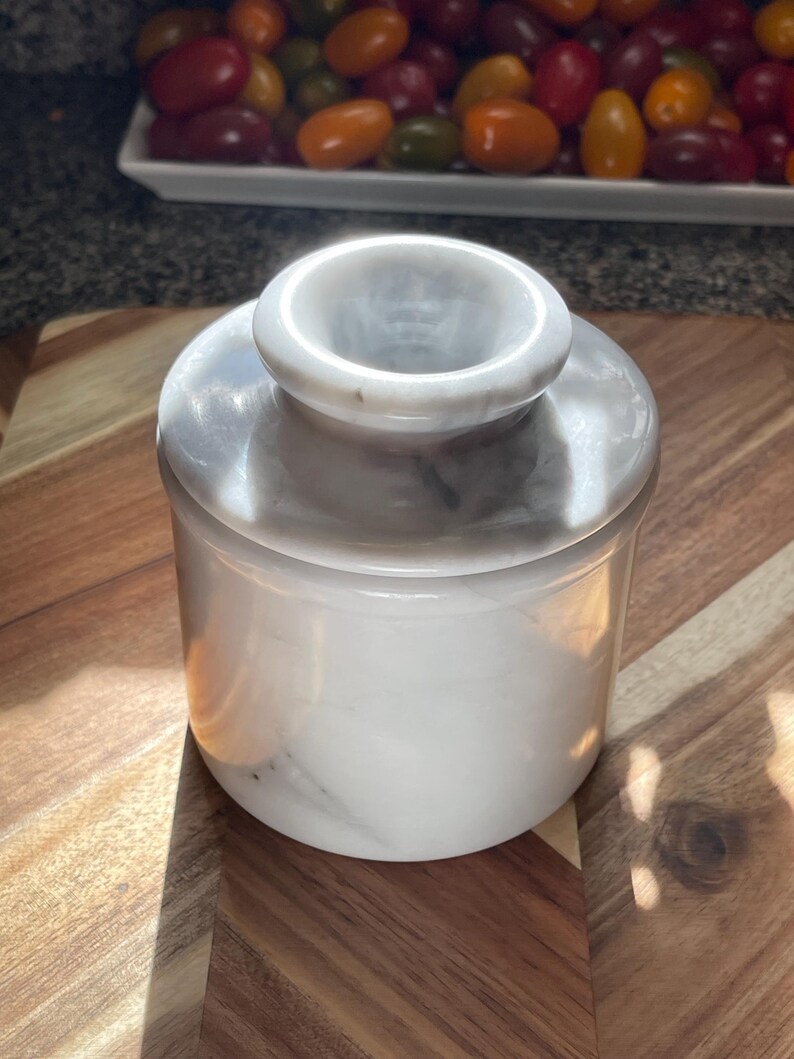 May include: A cylindrical white marble butter keeper with a smooth, polished finish and subtle grey veining. The container features a two-piece design with a deep base and a matching lid that has a central knob handle for easy removal and handling.