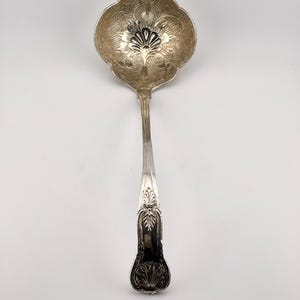 May include: A silver-toned metal ladle featuring a scalloped bowl with an intricate engraved floral pattern and a cross-hatched texture. The handle is long and slender, decorated with embossed leaf and shell motifs at the base and neck.