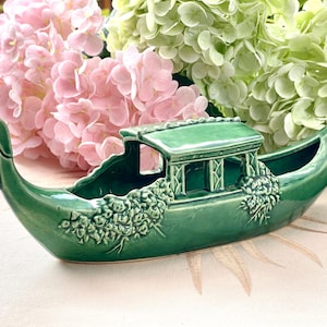 May include: A glossy emerald green ceramic gondola-shaped planter featuring a central roofed cabin structure and raised floral relief details on the hull. The vessel has a curved prow and stern, with an open interior space for holding small items.