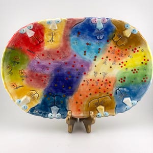 May include: An oval ceramic platter featuring a vibrant, multi-colored abstract design. The surface is divided into sections of red, yellow, blue, green, and orange, decorated with small painted animal faces, floral motifs, and geometric patterns.