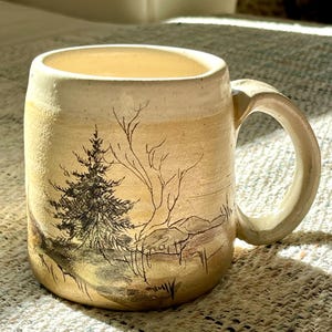 May include: A ceramic mug with a cream-colored glaze featuring a hand-drawn black ink illustration of a pine tree and a bare deciduous tree. The mug has a cylindrical body, a thick handle on the right side, and a textured, earthy finish.