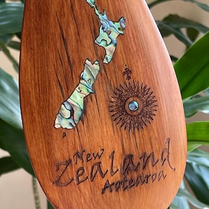 May include: A wooden paddle blade featuring an inlaid abalone shell map of New Zealand. The design includes a laser-engraved compass rose with a central abalone accent and the text New Zealand Aotearoa etched into the polished wood surface.