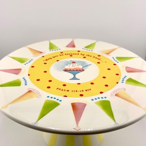 May include: A round ceramic cake stand with a white surface featuring a central yellow circle with red polka dots and an illustration of a sundae. The text reads, "May you be blessed by the Lord...." and "PSALM 115:15 NIV" with colorful party hat motifs.