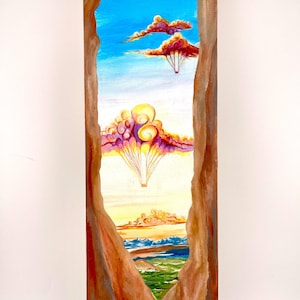 May include: A vertical painting framed by brown rock formations, depicting a bright blue sky with two hot air balloons. The balloons are shaped like stylized clouds in shades of orange, yellow, and purple, floating above a distant landscape.