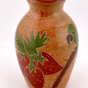 May include: A tan ceramic vase with a red rim featuring a hand-painted design of red strawberries with green leaves and a green parrot perched on a brown branch. The surface has a textured, speckled appearance with etched outlines.