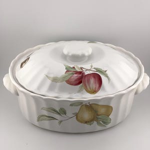 Op de afbeelding: Een witte keramische ovenschaal met een geribbelde rand en een bijpassend deksel. De buitenkant is voorzien van een geschilderde botanische illustratie van twee rode appels op het deksel en twee gele peren op de basis, met groene bladeren.