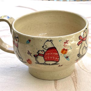 May include: A speckled cream-colored ceramic mug featuring a hand-painted illustration of polar bears wearing red patterned sweaters and scarves, accented with small gift boxes and foliage motifs, complete with a sturdy handle and a pedestal base.