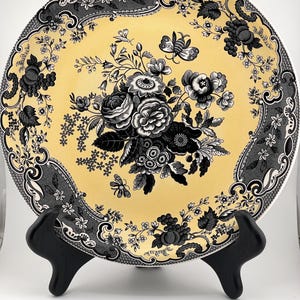May include: A round ceramic plate featuring a pale yellow center with a detailed black floral bouquet and insect illustrations. The border is decorated with a dark gray and black intricate pattern of leaves, vines, and small flowers.