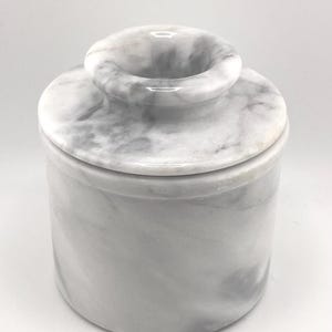 May include: A cylindrical white marble butter keeper with grey veining. The container features a smooth, polished finish and a removable lid with a central knob handle, designed for storing butter at room temperature in a kitchen setting.