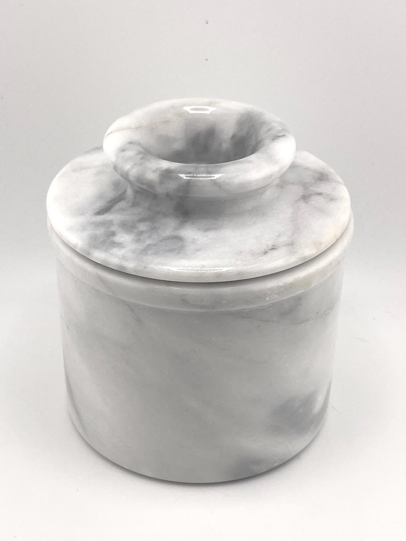 May include: A cylindrical white marble butter keeper with grey veining. The container features a smooth, polished finish and a removable lid with a central knob handle, designed for storing butter at room temperature in a kitchen setting.