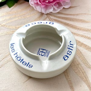 May include: A round white ceramic ashtray featuring blue text that reads "les hôtels astir" and "grèce" along the rim, with a central logo containing the letter "a" and the text "les hôtels astir" printed in the center of the basin.