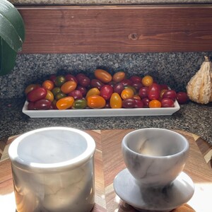May include: A cylindrical white marble container with a smooth finish and a matching white marble cup resting on a circular base, both featuring subtle grey veining throughout the stone surface, displayed on a wooden surface.