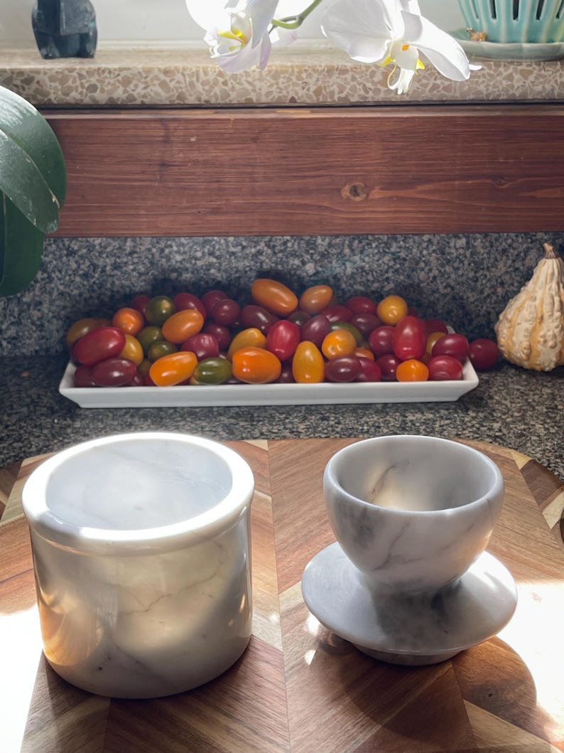 May include: A cylindrical white marble container with a smooth finish and a matching white marble cup resting on a circular base, both featuring subtle grey veining throughout the stone surface, displayed on a wooden surface.