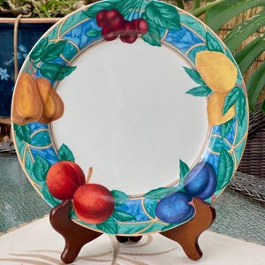 May include: A round ceramic plate featuring a white center and a decorative border. The border displays a blue and green stained-glass style pattern with illustrations of cherries, a lemon, plums, peaches, and pears arranged around the rim.