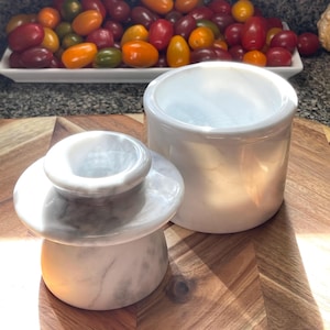 May include: A white marble butter keeper consisting of two pieces: a bell-shaped lid with a knob handle and a cylindrical base. The polished stone features natural grey veining throughout, providing a smooth, glossy finish to the kitchenware.