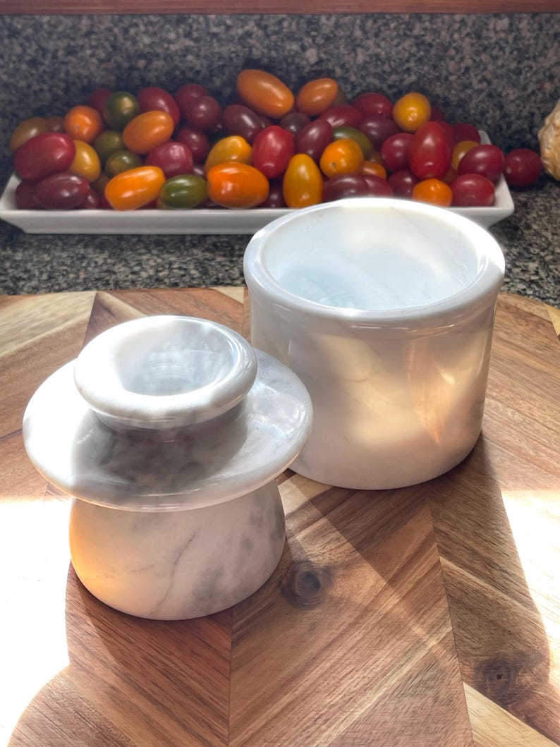 May include: A white marble butter keeper consisting of two pieces: a bell-shaped lid with a knob handle and a cylindrical base. The polished stone features natural grey veining throughout, providing a smooth, glossy finish to the kitchenware.