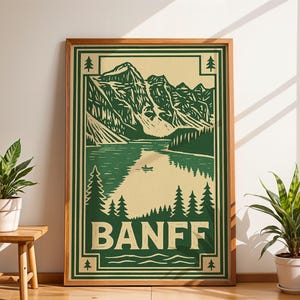 May include: A framed art print featuring a green and beige illustration of Banff National Park. The artwork depicts mountains, a lake with a small boat, and the word "BANFF" in bold letters. The frame is a natural wood color.