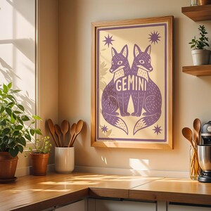 Puede incluir: Una impresi&oacute;n art&iacute;stica enmarcada con dos zorros morados y la palabra "GEMINI". La obra de arte est&aacute; sobre un fondo crema con acentos de estrellas. La impresi&oacute;n se exhibe en un entorno de cocina con estantes de madera y una cafetera.