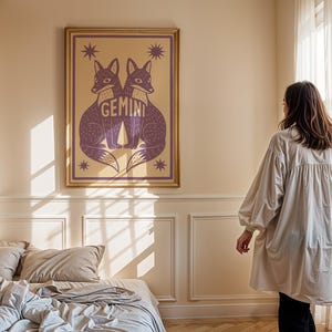Puede incluir: Una impresi&oacute;n art&iacute;stica enmarcada con dos zorros morados enfrentados, con la palabra "Gemini" en el centro. La obra de arte est&aacute; sobre un fondo beige con acentos de estrellas y un marco dorado. La impresi&oacute;n se exhibe en un dormitorio.