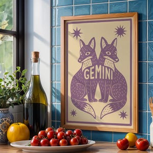 Puede incluir: Un cuadro enmarcado con dos zorros morados y la palabra "GEMINI". La obra tiene un fondo crema con detalles de estrellas, en un marco de madera. Se exhibe en una encimera con una botella de aceite de oliva, un jarr&oacute;n y un plato de tomates.