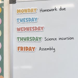 May include: A white magnetic whiteboard featuring five colourful die-cut labels for the days of the week from Monday to Friday. Handwritten black text next to the labels reads Homework due, Science incursion, and Assembly on the smooth surface.
