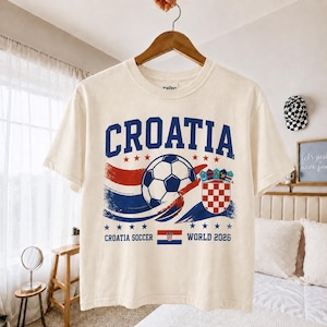 May include: A cream-coloured short-sleeve t-shirt featuring a blue and red graphic with the text CROATIA, a football, the Croatian coat of arms, and the text CROATIA SOCCER WORLD 2026 with a small Croatian flag centred below the football.