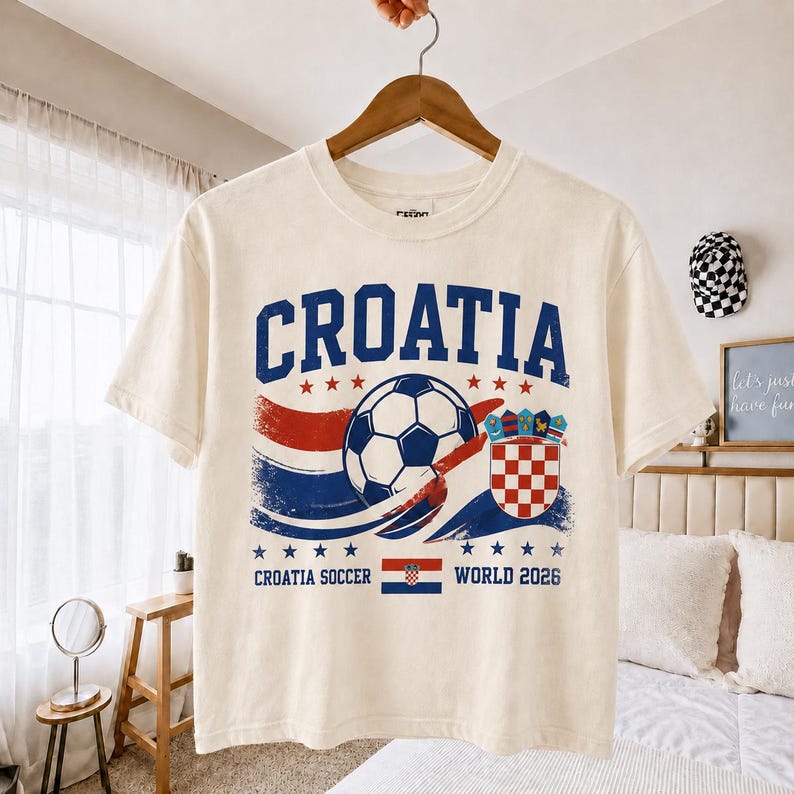 May include: A cream-coloured short-sleeve t-shirt featuring a blue and red graphic with the text CROATIA, a football, the Croatian coat of arms, and the text CROATIA SOCCER WORLD 2026 with a small Croatian flag centred below the football.