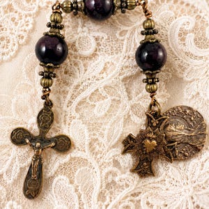 May include: A rosary featuring dark purple beads and ornate bronze-toned metalwork. The rosary includes two crosses, one with a crucifix and the other with a heart design, and a circular pendant. The beads are approximately 1 cm in diameter.