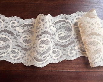 May include: Ivory-colored lace trim with a repeating floral and swirl pattern. The lace is folded over on the right side of the image, showing the intricate design. The trim is laid on a dark brown wooden surface.
