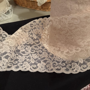 Puede incluir: Un rollo de tela de encaje color crema con un diseño floral. El encaje se muestra sobre una superficie negra, con una sección desenrollada, mostrando el intrincado patrón. Otros rollos de encaje son visibles en el fondo.