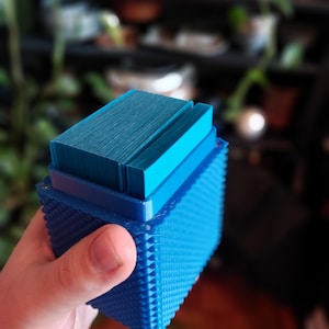 Può includere: Una scatola rettangolare blu, strutturata, con coperchio, tenuta in mano. La scatola presenta piccole sporgenze a forma di piramide sui lati. Il coperchio è di un blu più chiaro e ha una texture scanalata. La scatola sembra essere stampata in 3D.