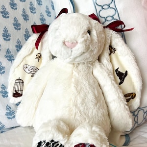 May include: A plush white bunny rabbit with burgundy bows on its ears. The inner ears feature a cream-colored fabric with whimsical illustrations. The bunny's feet display embroidered crests. The bunny is sitting on a white bed with patterned pillows in the background.