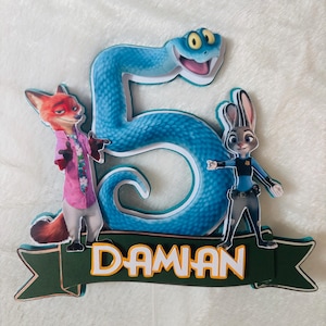 May include: A colorful cake topper featuring characters from Zootopia, including Nick Wilde and Judy Hopps, with a large blue number 5 and the name "Daman" in white letters on a green banner. The topper is designed for a birthday celebration.