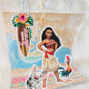 May include: A clear plastic tote bag featuring a Moana-themed graphic with Moana, Pua the pig, and Heihei the rooster. A surfboard-shaped tag attached by a white chain displays the text Melody Turns 5 with a hibiscus flower and tropical leaves.