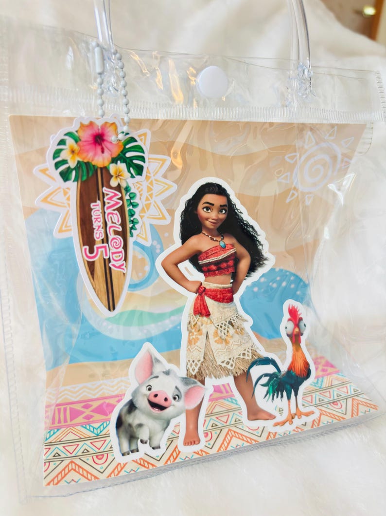 May include: A clear plastic tote bag featuring a Moana-themed graphic with Moana, Pua the pig, and Heihei the rooster. A surfboard-shaped tag attached by a white chain displays the text Melody Turns 5 with a hibiscus flower and tropical leaves.