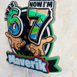 May include: A colorful cake topper with the text "I WAS 6, NOW I'M 7" in a graffiti style. The name "Maverik" is displayed on a banner. The design includes hands and geometric shapes, perfect for a birthday celebration.