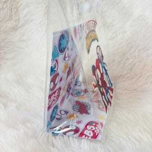 May include: Clear tote bag with transparent handles. The bag contains a card with colorful cartoon stickers and the text "OLIVER'S AMAZING". The stickers feature various superhero designs. The bag is made of clear plastic.