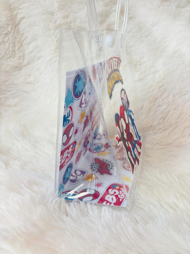 May include: Clear tote bag with transparent handles. The bag contains a card with colorful cartoon stickers and the text "OLIVER'S AMAZING". The stickers feature various superhero designs. The bag is made of clear plastic.