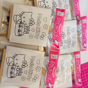May include: Several Hello Kitty coloring canvases with wooden frames are packaged in clear plastic. Each canvas features a black outline of Hello Kitty with the words "Hello Kitty" and decorative elements. Pink packaging with the name "Julianna" is also visible.
