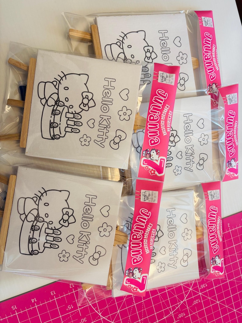 May include: Several Hello Kitty coloring canvases with wooden frames are packaged in clear plastic. Each canvas features a black outline of Hello Kitty with the words "Hello Kitty" and decorative elements. Pink packaging with the name "Julianna" is also visible.