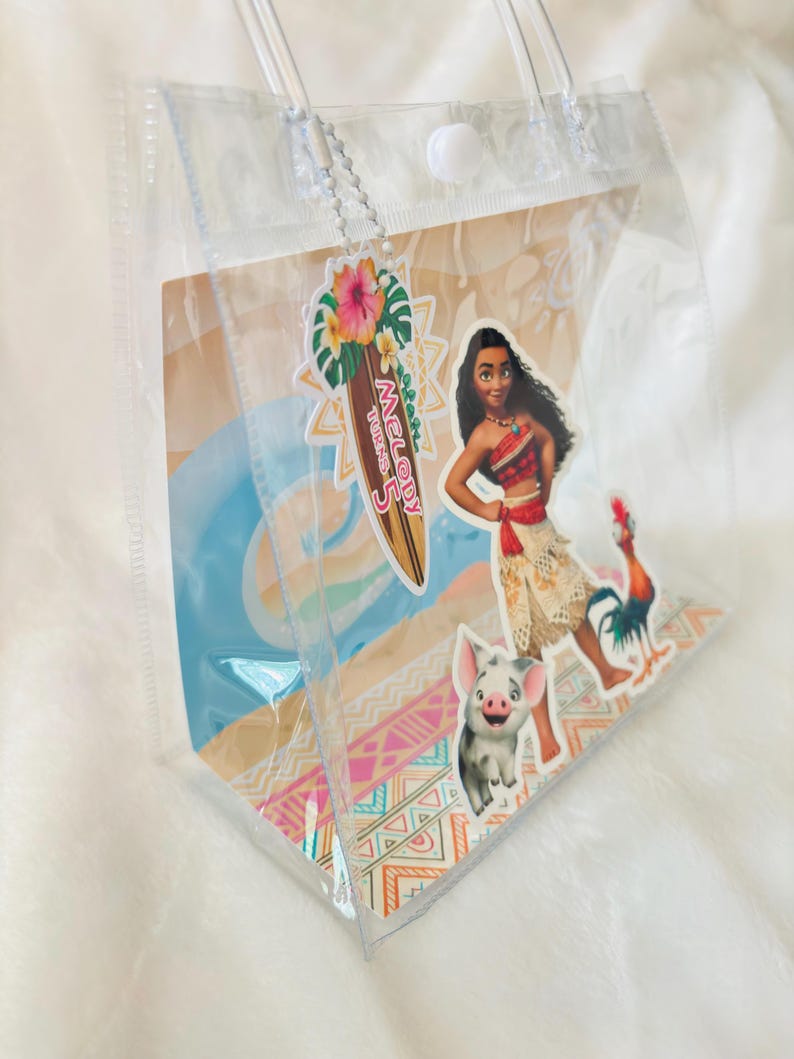 May include: A clear plastic tote bag with two transparent handles and a white snap closure. The bag features a decorative insert with Moana, Pua, and Heihei characters, plus a surfboard-shaped tag that reads Melody Turns 5.