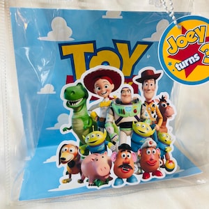 May include: A clear plastic bag featuring a blue background with white clouds and the word "Toy" in yellow. The bag displays various Toy Story characters, including Woody, Buzz Lightyear, and Jessie. A tag reads "Joey turns 3".