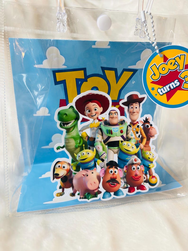 May include: A clear plastic bag featuring a blue background with white clouds and the word "Toy" in yellow. The bag displays various Toy Story characters, including Woody, Buzz Lightyear, and Jessie. A tag reads "Joey turns 3".