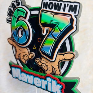 May include: A colorful cake topper with the text "I WAS 6 NOW I'M 7" in a graffiti style. The design includes the name "Maverik" at the bottom, with a black, green, and red color scheme. The numbers 6 and 7 are in a vibrant green and blue.