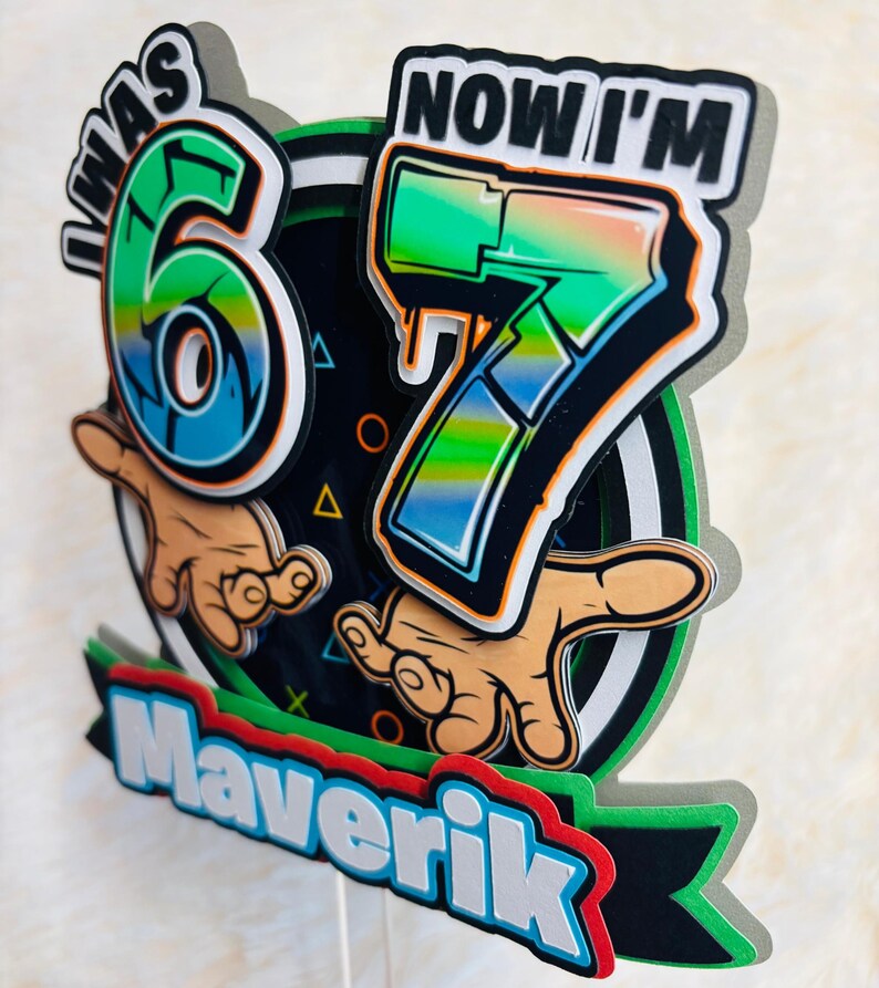 May include: A colorful cake topper with the text "I WAS 6 NOW I'M 7" in a graffiti style. The design includes the name "Maverik" at the bottom, with a black, green, and red color scheme. The numbers 6 and 7 are in a vibrant green and blue.