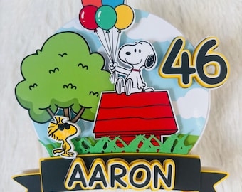 May include: A Snoopy-themed cake topper featuring Snoopy holding balloons, a red doghouse, a green tree, and the number 46. The name "Aaron" is displayed on a banner. The background is a blue sky with clouds.