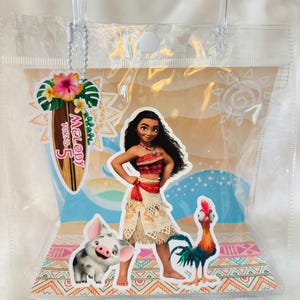 May include: A clear plastic tote bag featuring a Moana-themed insert with Moana, Pua the pig, and Heihei the rooster. A surfboard-shaped tag attached by a silver chain reads Melody Turns 5, decorated with a hibiscus flower and tropical leaves.